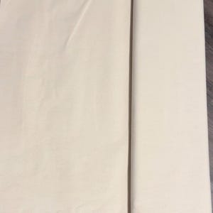 May include: A folded and rolled piece of light beige fabric. The fabric is smooth and appears to be a natural material. The image shows the fabric partially rolled, with a visible crease where it is folded.