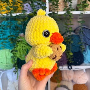 Puede incluir: Un patito amarillo de ganchillo hecho a mano con patas y pico naranjas, sosteniendo un peque&ntilde;o osito de peluche marr&oacute;n. El patito tiene ojos negros y una textura esponjosa. Un juguete suave y lindo.