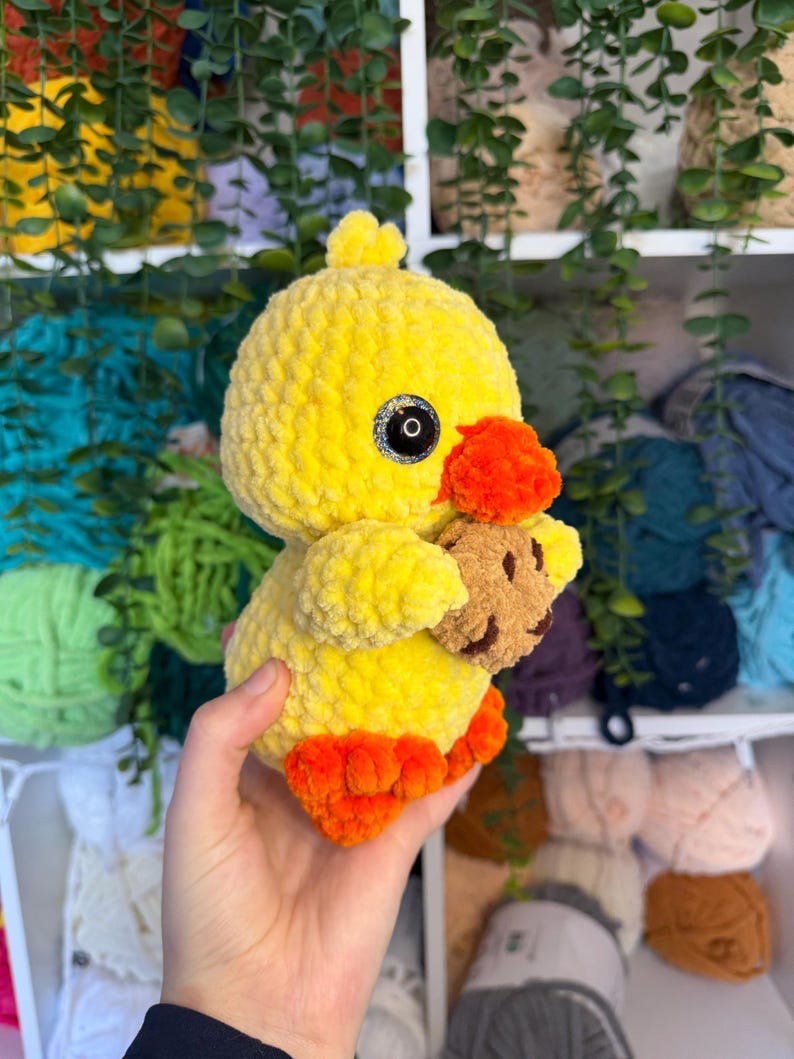 Puede incluir: Un patito amarillo de ganchillo hecho a mano con patas y pico naranjas, sosteniendo un peque&ntilde;o osito de peluche marr&oacute;n. El patito tiene ojos negros y una textura esponjosa. Un juguete suave y lindo.