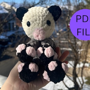 Puede incluir: Un juguete de peluche de crochet que se asemeja a un panda con pelaje blanco y negro, nariz rosa y ojos negros. El juguete tiene almohadillas de patas rosas y negras.