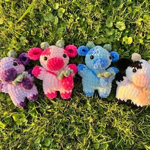 Peut inclure: Quatre vaches en peluche crochetées de différentes couleurs : violet, rose, bleu et noir et blanc. Les vaches sont assises sur un lit d’herbe verte.