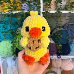 Puede incluir: Un pato amarillo hecho a mano con pies y pico naranjas, sosteniendo una galleta marr&oacute;n con una cara sonriente. El pato tiene ojos negros y est&aacute; hecho de hilo suave y texturizado. Un juguete de peluche lindo y caprichoso.
