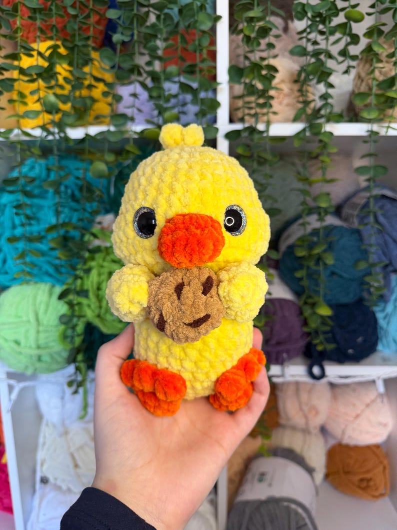 Puede incluir: Un pato amarillo hecho a mano con pies y pico naranjas, sosteniendo una galleta marr&oacute;n con una cara sonriente. El pato tiene ojos negros y est&aacute; hecho de hilo suave y texturizado. Un juguete de peluche lindo y caprichoso.