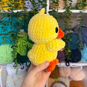 Puede incluir: Un pato amarillo de peluche hecho a mano, con patas y pico naranjas. El pato tiene ojos negros y una superficie texturizada y difusa. El juguete se sostiene en la mano de una persona, con un fondo de hilo y vegetaci&oacute;n.