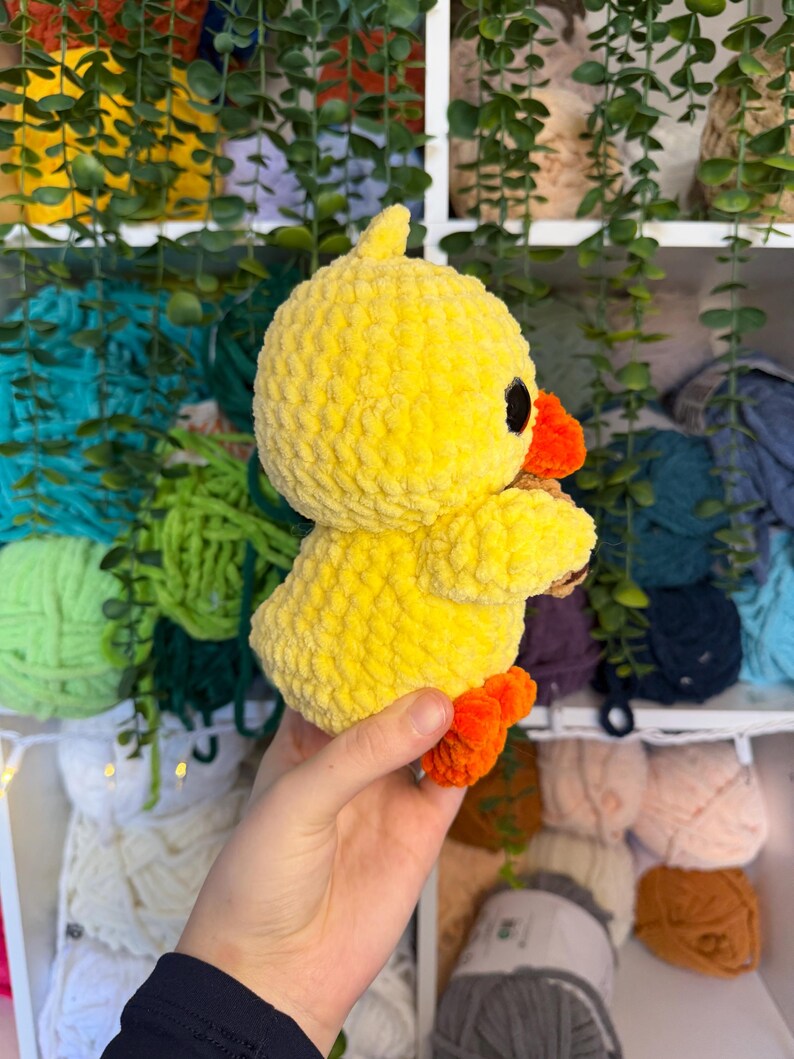 Puede incluir: Un pato amarillo de peluche hecho a mano, con patas y pico naranjas. El pato tiene ojos negros y una superficie texturizada y difusa. El juguete se sostiene en la mano de una persona, con un fondo de hilo y vegetaci&oacute;n.