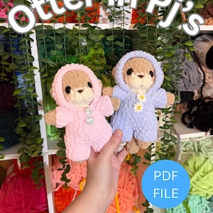 May include: Two plush crochet otter toys, one wearing a pink hooded onesie with two buttons and the other in a light blue hooded onesie with two daisy flower accents. The text Otter in Pj's and PDF File Crochet Pattern is visible.