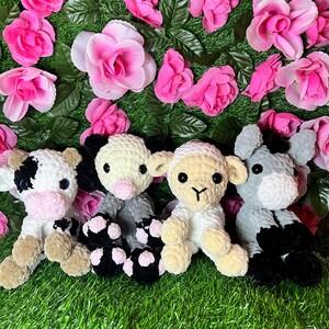 May include: Four crocheted stuffed animals: a cow, a pig, a lamb, and a donkey. The animals are sitting on a green grassy surface with pink flowers.