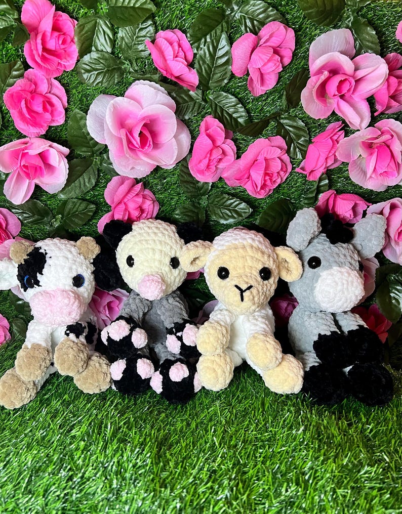 May include: Four crocheted stuffed animals: a cow, a pig, a lamb, and a donkey. The animals are sitting on a green grassy surface with pink flowers.