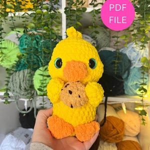 Puede incluir: Un pato amarillo hecho a mano con ganchillo sosteniendo una galleta. El pato tiene patas y pico naranjas, con grandes ojos azules. La imagen incluye un c&iacute;rculo rosa con el texto "PDF FILE".