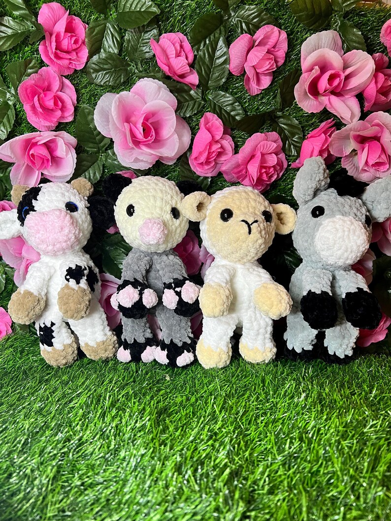 May include: Four crocheted stuffed animals: a cow, a pig, a lamb, and a donkey. The animals are all white with black spots or stripes, and they have pink noses and eyes. They are sitting on a green grassy surface.