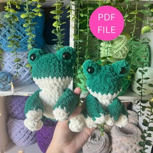 May include: Two handmade crochet frog plushies. The frogs are green and white with black eyes. The image also shows a pink circle with the text "PDF FILE". The background includes yarn and greenery.