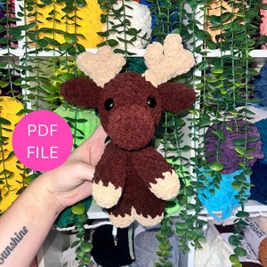 May include: A handmade, plush moose toy in shades of brown and beige. The moose has large, beige antlers, black eyes, and beige hooves. The text "PDF FILE" is in a pink circle in the image. The background includes shelves with yarn and hanging greenery.