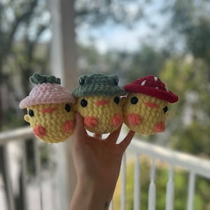 May include: Three crocheted plush toys shaped like chicks, each wearing a different hat. The first chick is wearing a pink and white mushroom hat, the second chick is wearing a green frog hat, and the third chick is wearing a red mushroom hat.