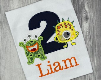 May include: White t-shirt with a navy blue number 2 and two embroidered monster characters. The name "Liam" is embroidered in orange below the monsters. The monsters are green and yellow with various details.