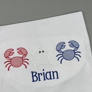 May include: White fabric with three embroidered crabs in red, white, and blue. The name "Brian" is embroidered in navy blue below the crabs. The design is likely for a child's item.