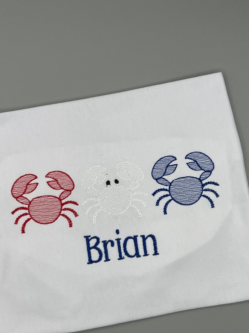 May include: White fabric with three embroidered crabs in red, white, and blue. The name "Brian" is embroidered in navy blue below the crabs. The design is likely for a child's item.