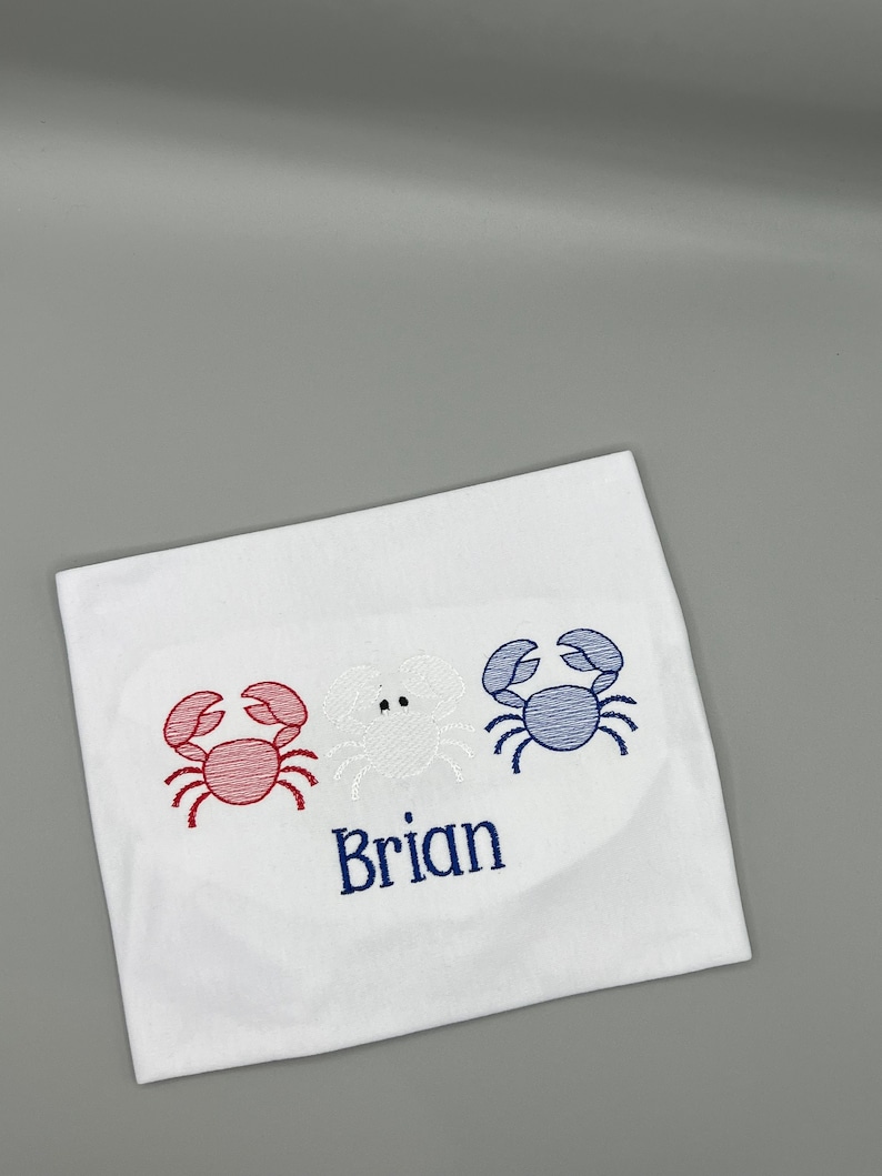 May include: White fabric with embroidered crabs in red, white, and blue. The name "Brian" is embroidered in blue below the crabs. The design is centered on the fabric.