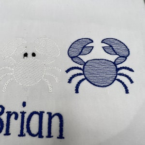 May include: White fabric with embroidered crab designs. One crab is white, the other is blue. The name "Brian" is embroidered in blue below the crabs. The design is likely for a nautical-themed item.