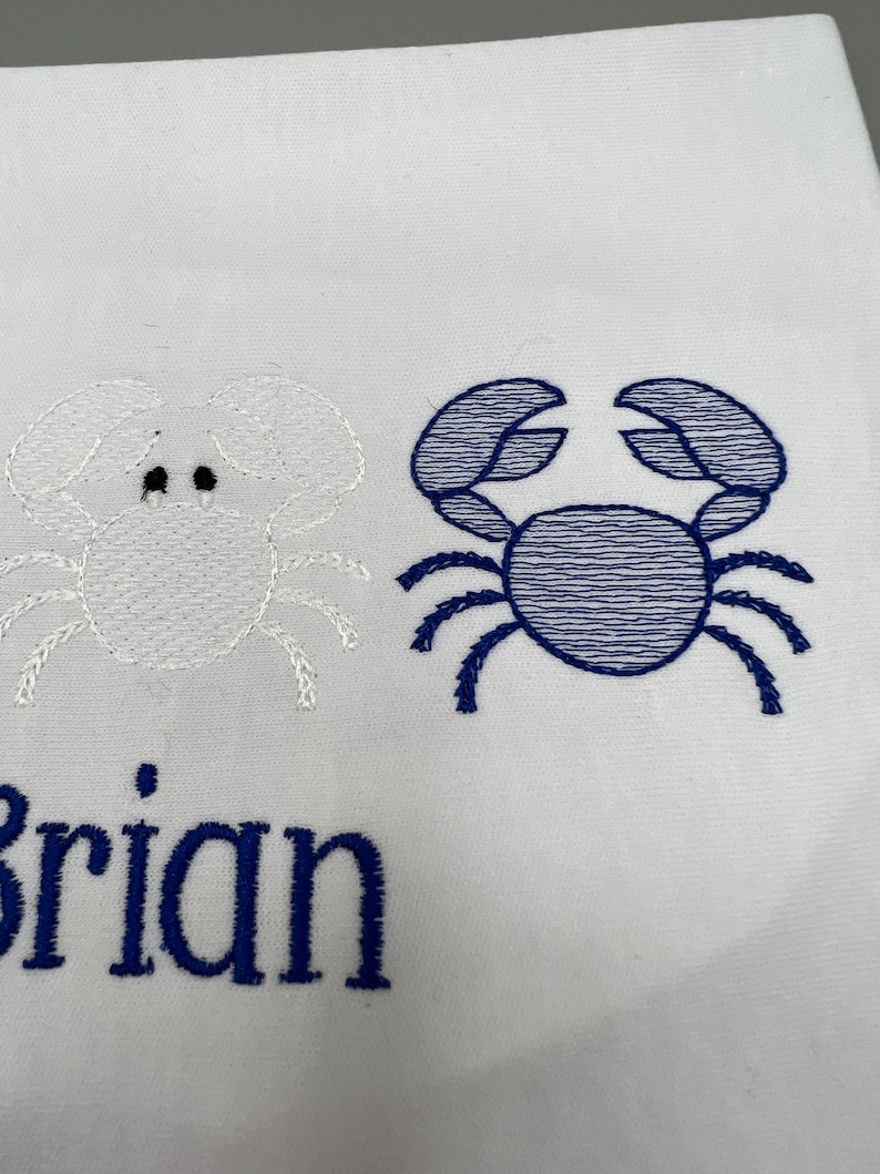 May include: White fabric with embroidered crab designs. One crab is white, the other is blue. The name "Brian" is embroidered in blue below the crabs. The design is likely for a nautical-themed item.