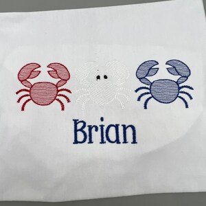 May include: White fabric with embroidered crabs in red, white, and blue. The name "Brian" is embroidered in blue below the crabs. The design is nautical themed.