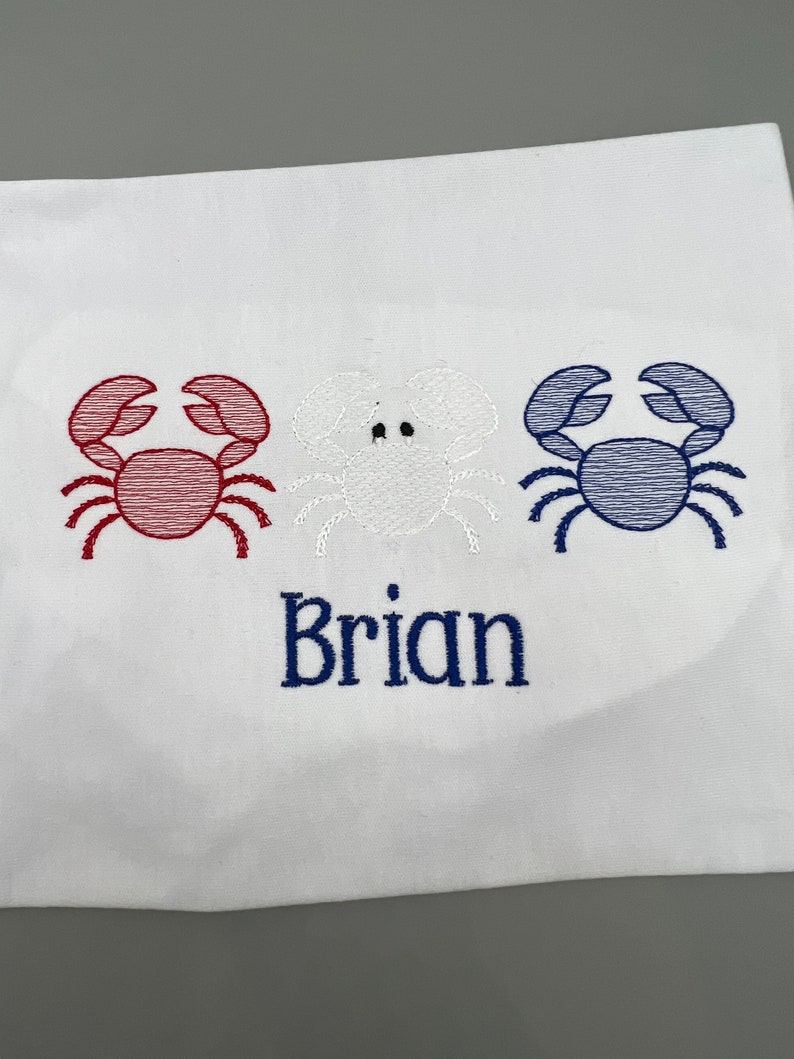 May include: White fabric with embroidered crabs in red, white, and blue. The name "Brian" is embroidered in blue below the crabs. The design is nautical themed.