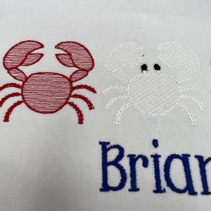 May include: White fabric embroidered with two crab designs. One crab is red, the other is white. The name "Brian" is embroidered in blue below the crabs.