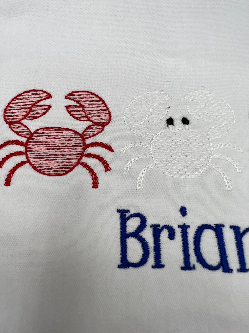May include: White fabric embroidered with two crab designs. One crab is red, the other is white. The name "Brian" is embroidered in blue below the crabs.