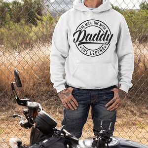 May include: A white hooded sweatshirt with a black circular graphic that reads "The Man The Myth Daddy The Legend". A person is wearing the sweatshirt and standing in front of a black motorcycle.