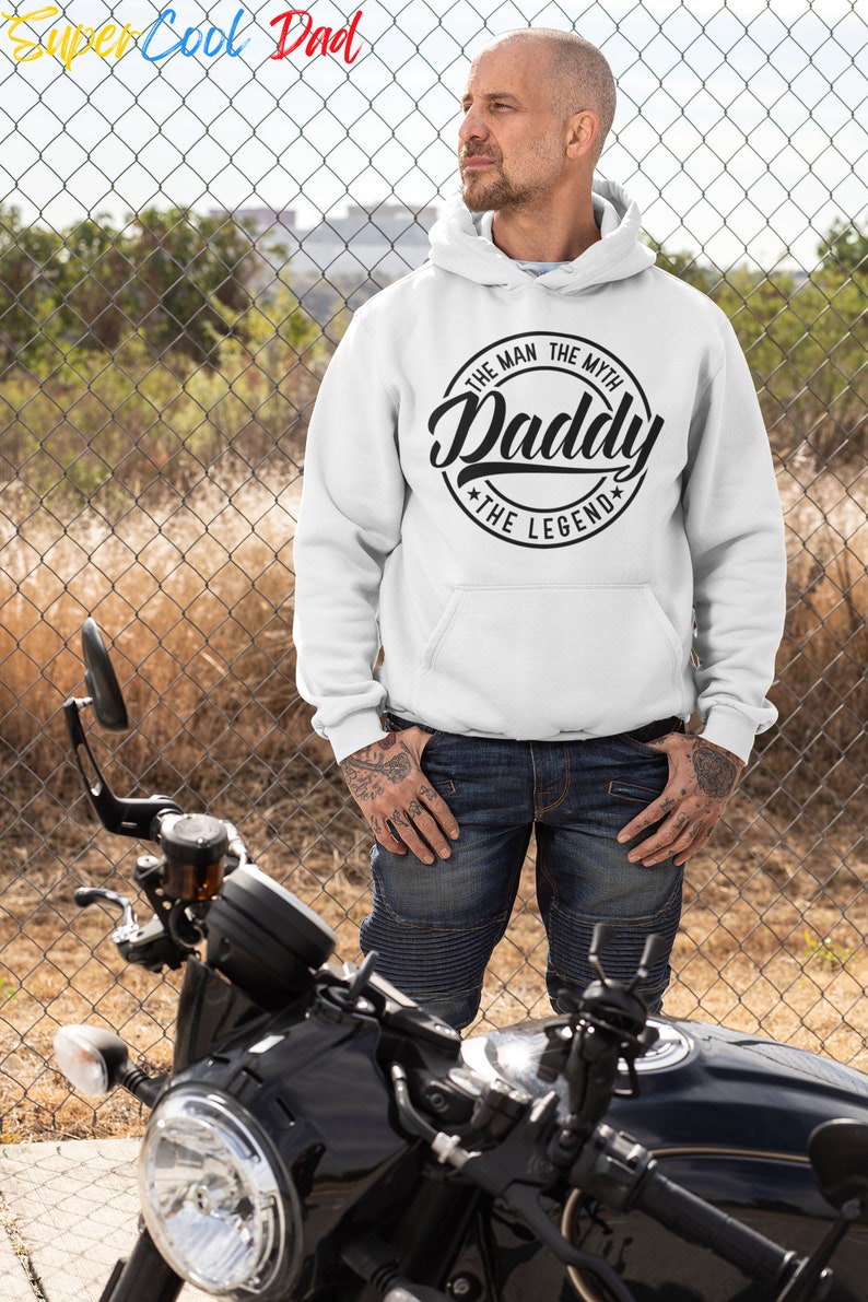 May include: A white hooded sweatshirt with a black circular graphic that reads "The Man The Myth Daddy The Legend". A person is wearing the sweatshirt and standing in front of a black motorcycle.