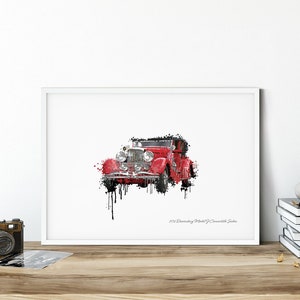 May include: A framed print of a red vintage Duesenberg Model J Convertible Sedan car. The artwork has a watercolor effect with black paint drips. The print is displayed on a wooden surface with a camera and other decorative items.