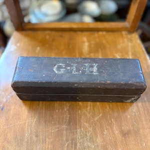 May include: A rectangular, dark brown wooden box with the initials "G.L.H" etched on the top. The box appears aged, with visible wear and tear. The box is resting on a wooden surface.