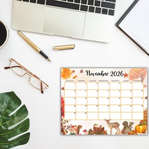 Puede incluir: Un calendario de escritorio para noviembre de 2026 con tem&aacute;tica oto&ntilde;al. El calendario presenta un dise&ntilde;o de cuadr&iacute;cula con un borde decorativo de follaje oto&ntilde;al, calabazas, ardillas y un ciervo. Est&aacute; rodeado por un port&aacute;til, un bol&iacute;grafo, gafas y una taza de caf&eacute;.