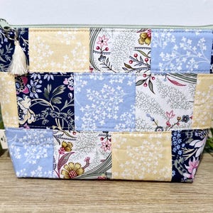 May include: A patchwork cosmetic bag with a zip closure and a tassel. The bag features floral patterns in navy, light blue, and yellow. The bag is approximately 20 cm wide and 15 cm tall.
