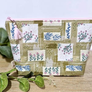 May include: A floral print cosmetic bag with a pink zip and tassel. The bag features a repeating pattern of white squares with pink flowers and blue leaves on a tan background. The bag is made of fabric and is designed to hold makeup or other small items.