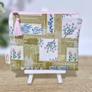 May include: A small, square, zipped pouch featuring a floral and leaf design in green, white, and pink. It has a pink zip and a pink tassel. The pouch is displayed on a small white easel.