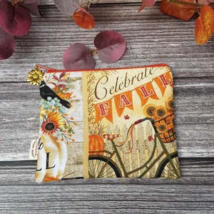 May include: A small zippered pouch with a fall-themed design. The pouch features a bicycle with a basket of sunflowers, a bird, and the text "Celebrate Fall".