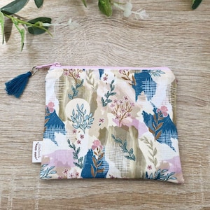 May include: A rectangular, floral-patterned pouch with a pink zipper and a blue tassel. The fabric features a design with blue, pink, and beige botanical elements. A small white tag with text is attached to the side.