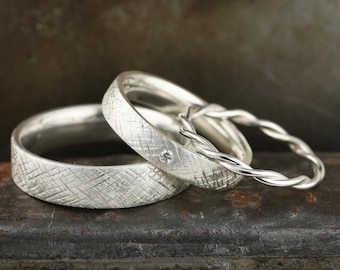 May include: Three silver wedding bands, two with a textured finish and one with a braided design. One of the textured bands has a small diamond.