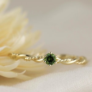 Könnte beinhalten: Ein goldener Ring mit einem gedrehten Band-Design und einem runden, grünen Edelstein in der Mitte. Der Ring ist vor einem weichen, neutralen Hintergrund platziert, mit einer unscharfen Blume im Vordergrund.