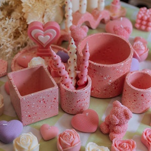 Op de afbeelding: Een assortiment van roze en witte hartvormige kaarsen, sommige met een gespikkelde afwerking, in verschillende maten en vormen. De kaarsen zijn gerangschikt op een tafel met een roze en wit geruit tafelkleed.