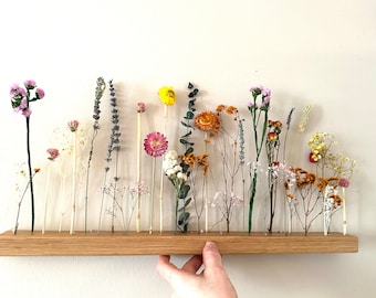 Può includere: Uno scaffale in legno con fiori secchi di vari colori, tra cui rosa, giallo, arancione e bianco. I fiori sono disposti in fila e lo scaffale è montato su una parete bianca.