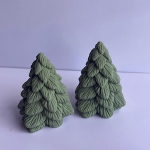 Könnte beinhalten: Zwei kleine, grüne, dekorative Weihnachtsbaumfiguren. Die Bäume haben ein strukturiertes, geschichtetes Design, wobei jede Schicht Nadeln ähnelt. Die Figuren stehen vor einem schlichten weißen Hintergrund.