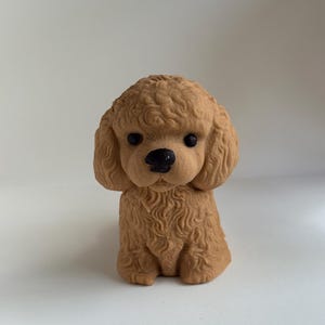 Może przedstawiać: Mała, brązowa figurka pudla z teksturowaną, kręconą sierścią. Pies ma czarny nos i czarne oczy i siedzi wyprostowany. Figurka jest prawdopodobnie wykonana z litego materiału i jest przedmiotem dekoracyjnym.