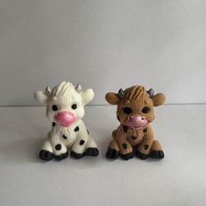 Puede incluir: Dos figuras de vaca en miniatura. Una es blanca con manchas negras, la otra es marrón con manchas negras. Ambas tienen narices rosas, cuernos pequeños y están sentadas.