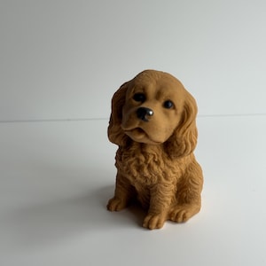 May include: A small, tan-colored figurine of a Cavalier King Charles Spaniel puppy. The puppy is sitting upright with its head slightly tilted, showcasing its floppy ears and detailed fur texture. The figurine has black eyes and a small, black nose.
