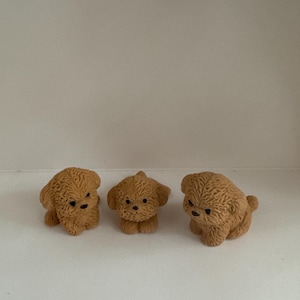 Może przedstawiać: Trzy małe, brązowe, puszyste figurki psów z czarnymi oczami i nosami. Psy są w różnych pozach, jeden siedzi, jeden stoi, a jeden jest zwinięty. Figurki są prawdopodobnie wykonane z żywicy lub podobnego materiału.