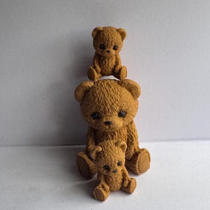 May include: Three brown teddy bear figurines stacked on top of each other. The largest bear sits at the bottom, with a medium-sized bear on its head, and a small bear on top. The bears have dark eyes and are made of a textured material.