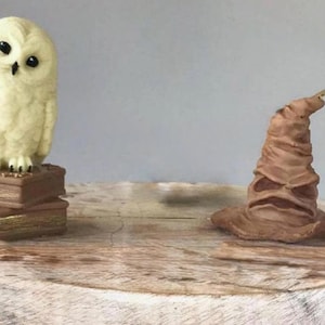 Könnte beinhalten: Eine cremefarbene Eulenfigur thront auf zwei braunen Büchern mit goldfarbenen Kanten. Daneben liegt eine braune Sprechender Hut-Figur auf einer Holzoberfläche. Die Figuren stammen wahrscheinlich aus einer Fantasy-Serie.