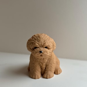 Puede incluir: Una figura de cachorro sentado de color marrón claro con un pelaje rizado y texturizado. El perro pequeño tiene ojos oscuros y una nariz negra. La figura está sobre una superficie blanca.