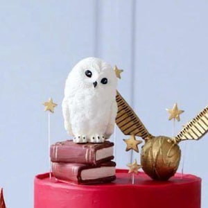 May include: A red cake decorated with a white owl figurine, two stacked books, a golden snitch, and star-shaped accents. The cake is likely themed for a special occasion.